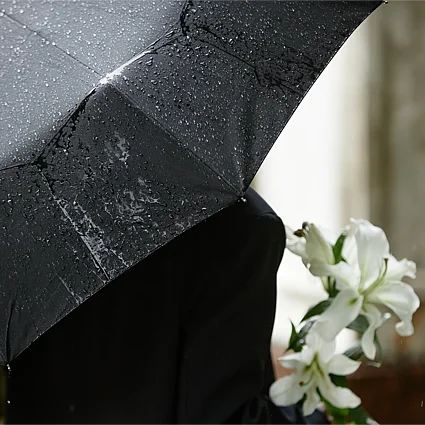 Person mit schwarzem Regenschirm und weißen Blumen im Regen – Symbol für Vorsorge und finanzielle Absicherung im Todesfall.