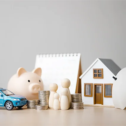 Modelle von Haus, Auto und Familie mit Schutzsymbolen als Darstellung von Sach- und Unfallversicherung sowie umfassender Absicherung im Alltag.