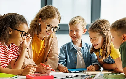 Lehrerin unterstützt lachende Kinder beim gemeinsamen Lernen im Klassenzimmer.